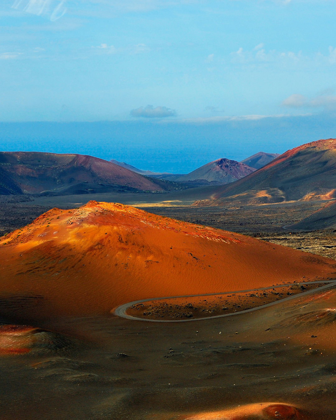 A scenic landscape photograph of a winding road cutting through orange-brown volcanic hills under a blue sky.