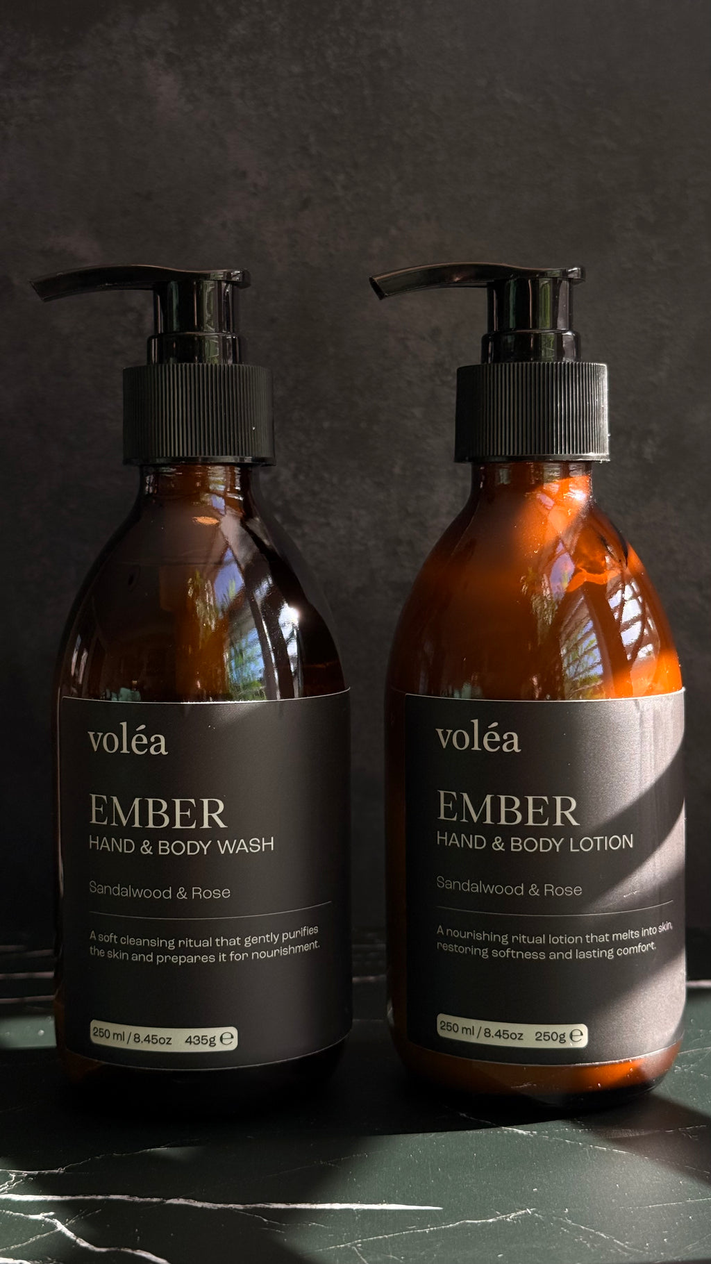 Two amber glass pump bottles of voléa Ember Hand & Body Wash and Hand & Body Lotion on a dark marble surface against a dark textured background.