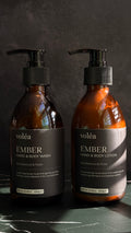Two amber glass pump bottles of voléa Ember Hand & Body Wash and Hand & Body Lotion on a dark marble surface against a dark textured background.