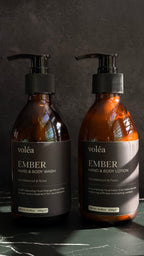 Two amber glass pump bottles of voléa Ember Hand & Body Wash and Hand & Body Lotion on a dark marble surface against a dark textured background.