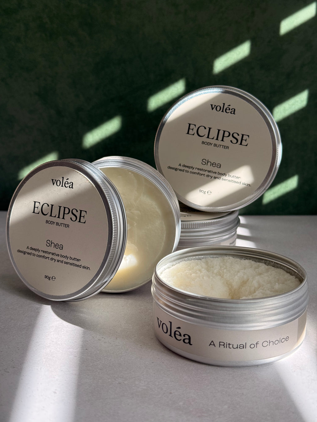 Volea Eclipse Shea Body Butter in a round silver tin, with one tin open to show the creamy off-white balm inside and another tin showing its lid label.