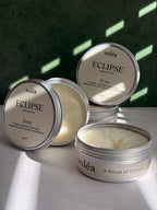 Volea Eclipse Shea Body Butter in a round silver tin, with one tin open to show the creamy off-white balm inside and another tin showing its lid label.