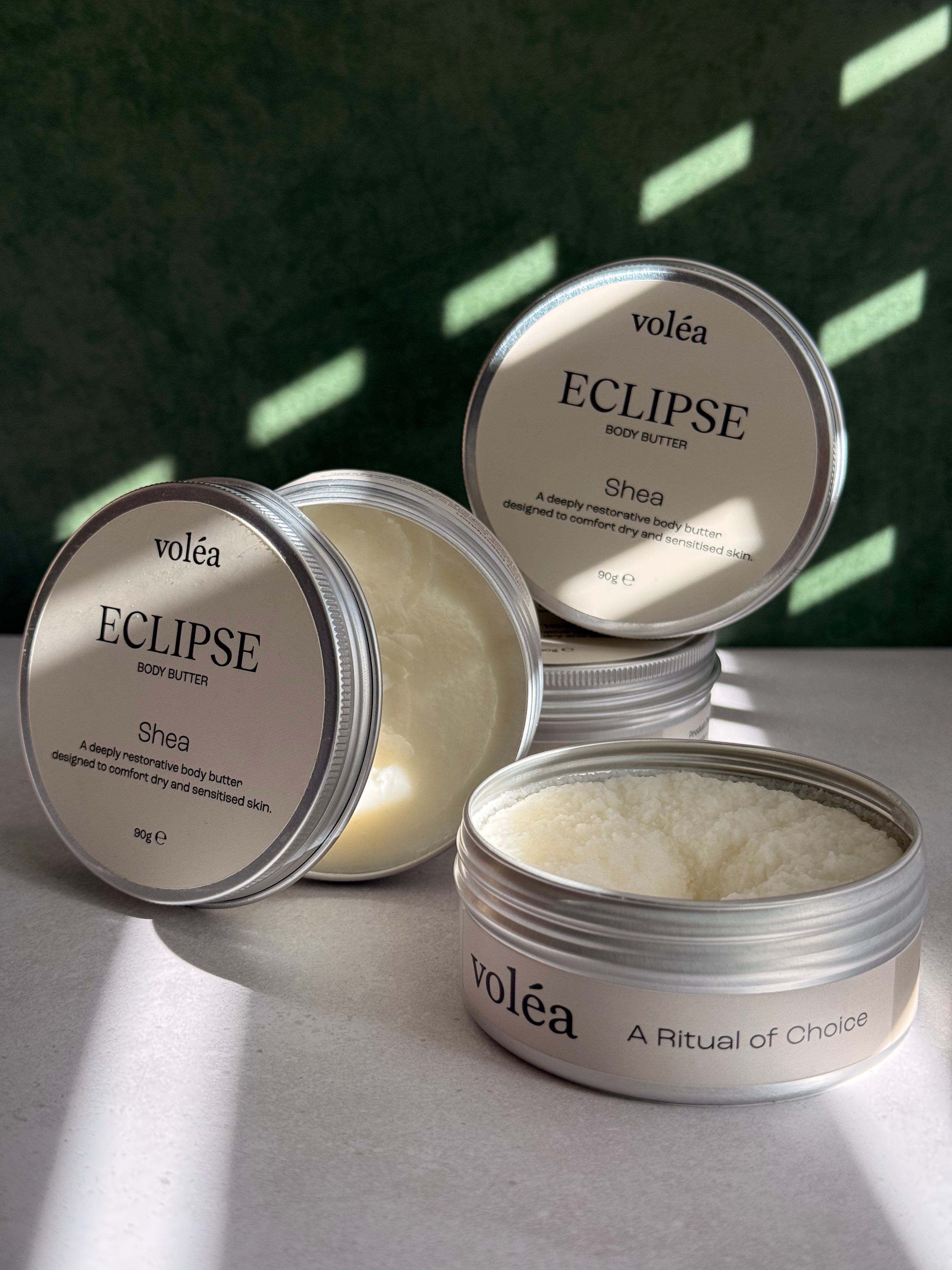 Volea Eclipse Shea Body Butter in a round silver tin, with one tin open to show the creamy off-white balm inside and another tin showing its lid label.