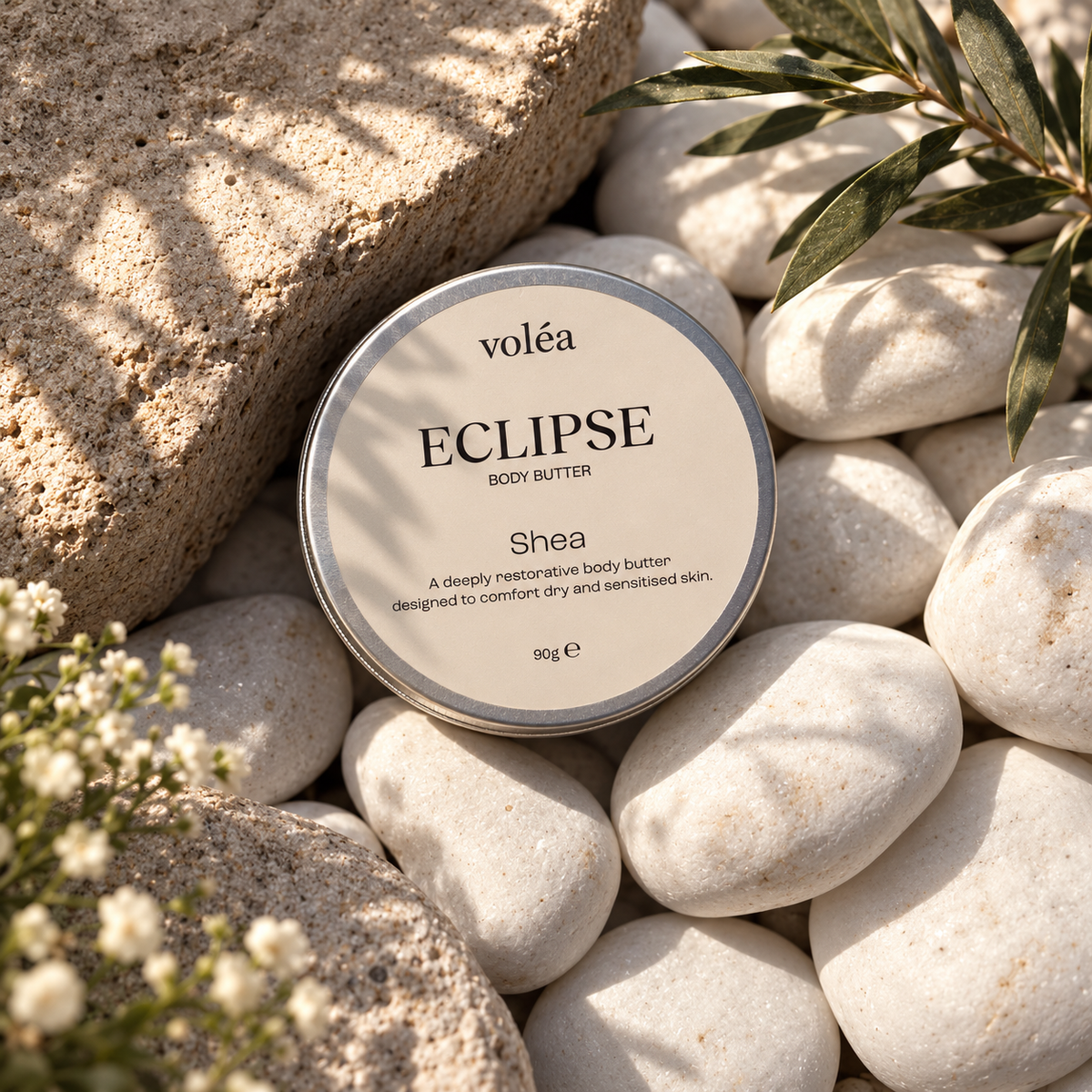 A round tin of voléa Eclipse Shea body butter sits on smooth white stones with natural sunlight and shadows, surrounded by textured rocks and green leaves.