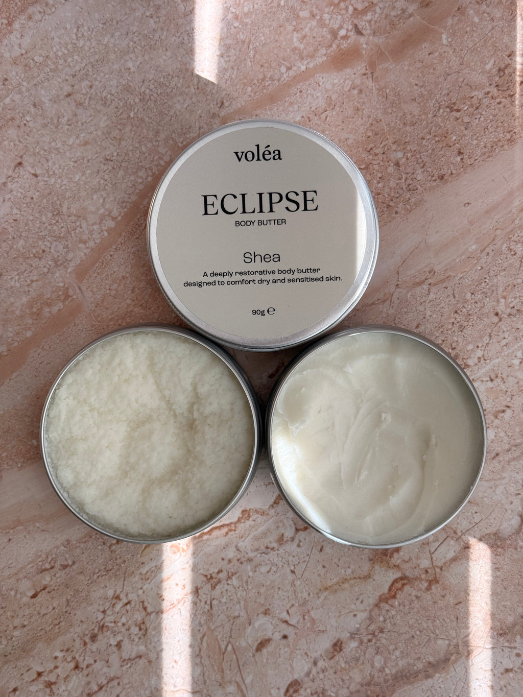 Volea Eclipse Shea Body Butter in a round silver tin, with one tin open to show the creamy off-white balm inside and another tin showing its lid label.