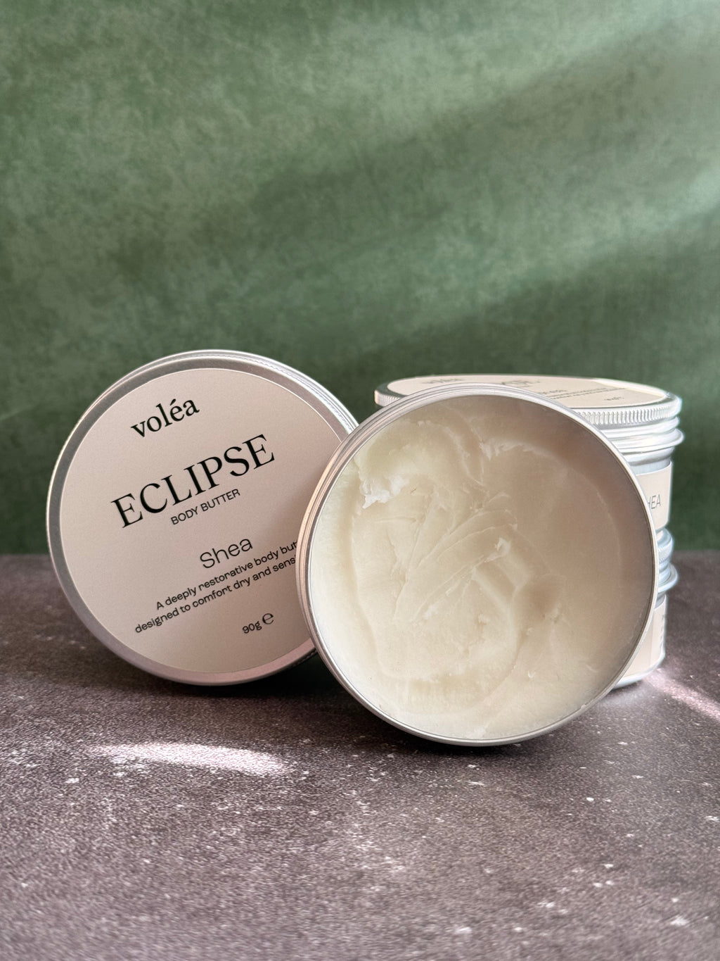 Volea Eclipse Shea Body Butter in a round silver tin, with one tin open to show the creamy off-white balm inside and another tin showing its lid label.