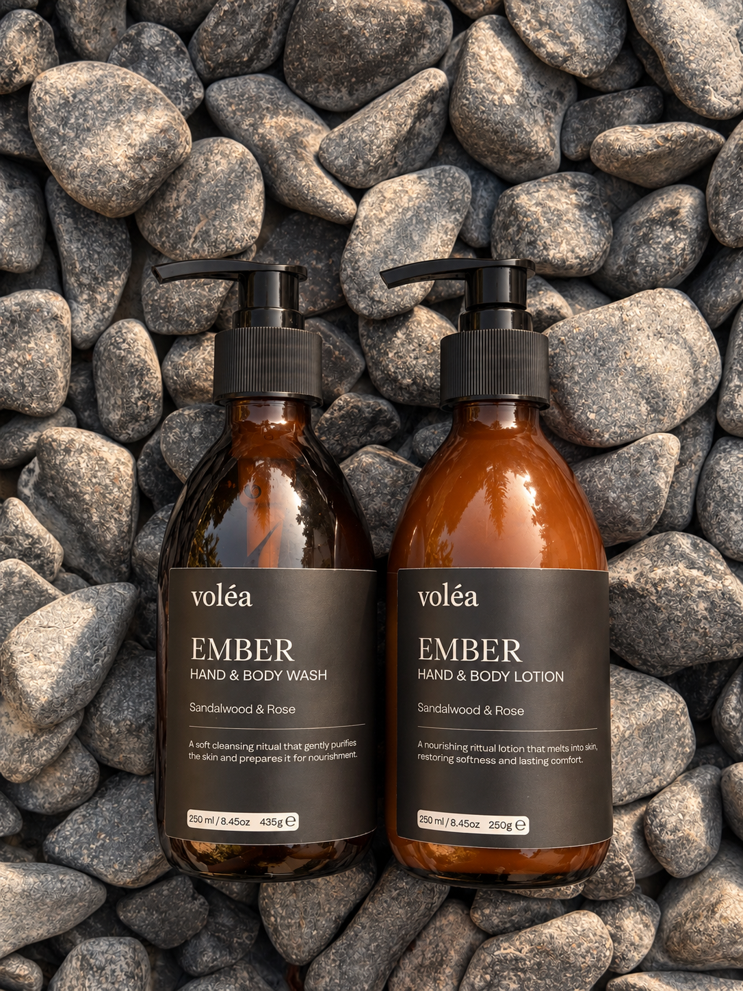 Two amber glass pump bottles of voléa EMBER Hand & Body Wash and EMBER Hand & Body Lotion, arranged side-by-side on a background of gray river stones.