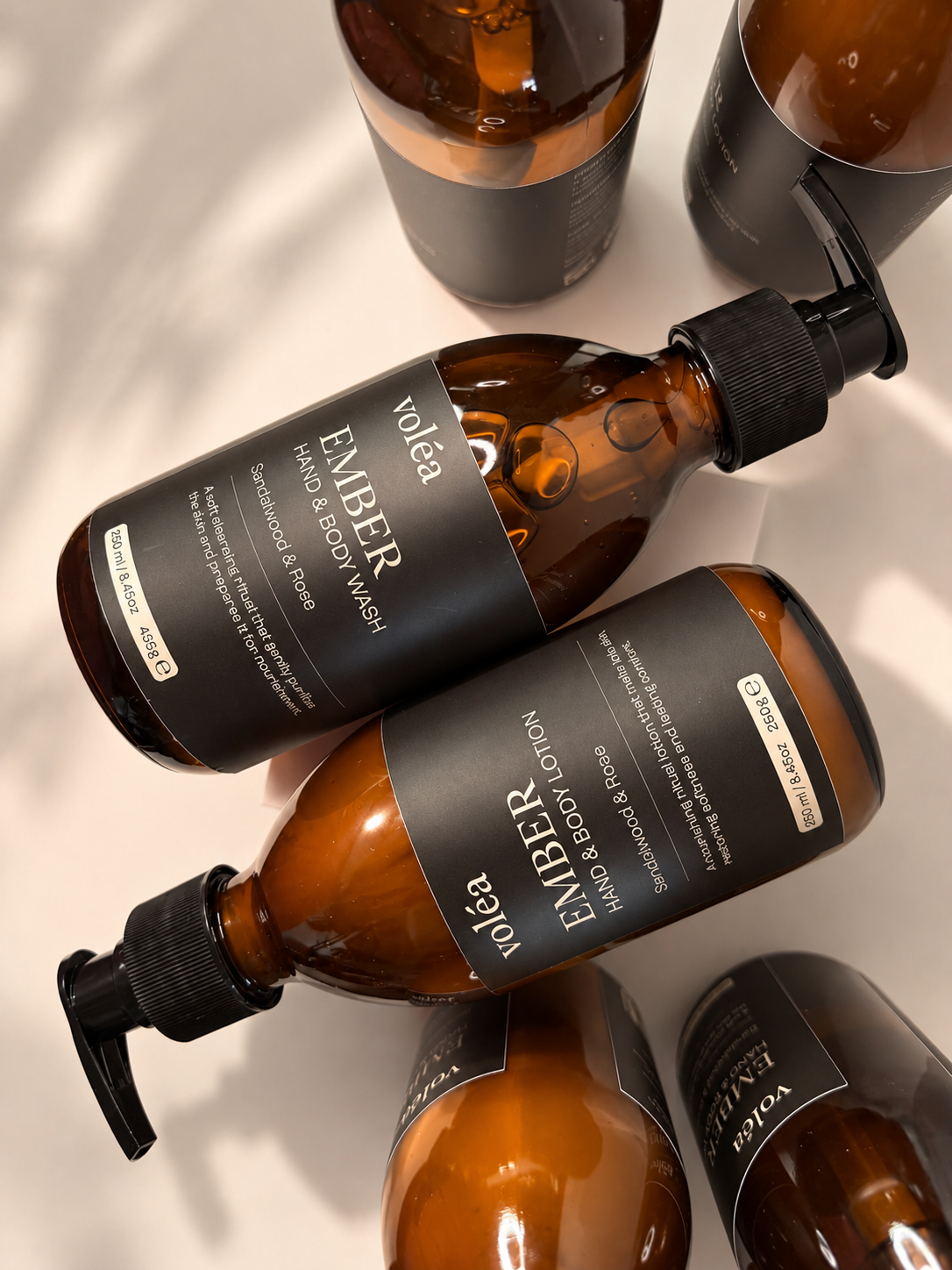 Several amber glass bottles with black pump and flip-top caps and black labels are arranged on a light surface. The visible label text includes the brand 'voka' and the line name 'EMBER'.