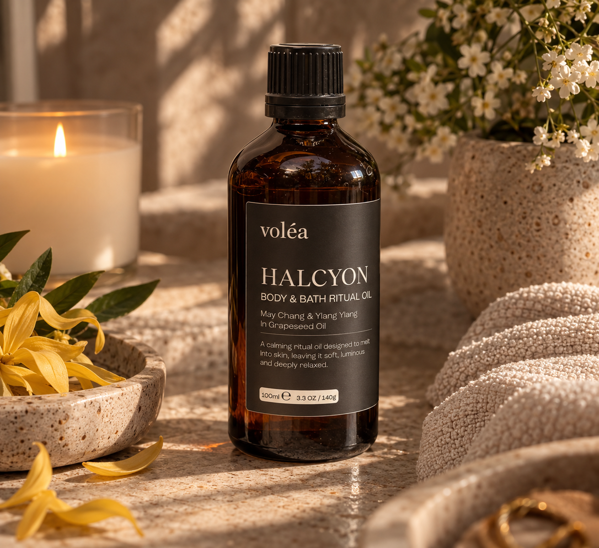 A bottle of voléa Halcyon body and bath ritual oil stands on a stone surface in a warm, styled setting with a lit candle, flowers, and folded towels. The amber glass bottle has a black cap and a black label with white text.