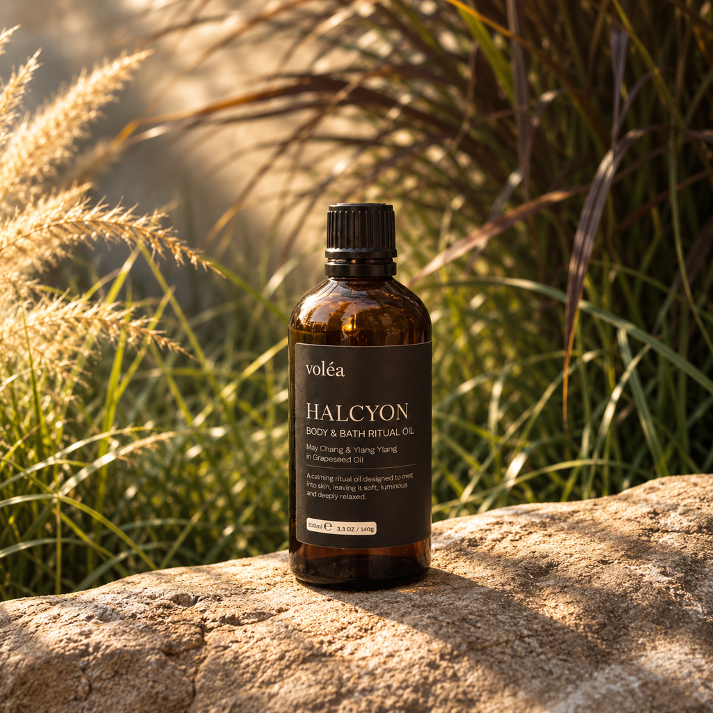 A bottle of voléa Halcyon Body & Bath Ritual Oil placed on a textured stone with a blurred natural background of green and beige grasses.