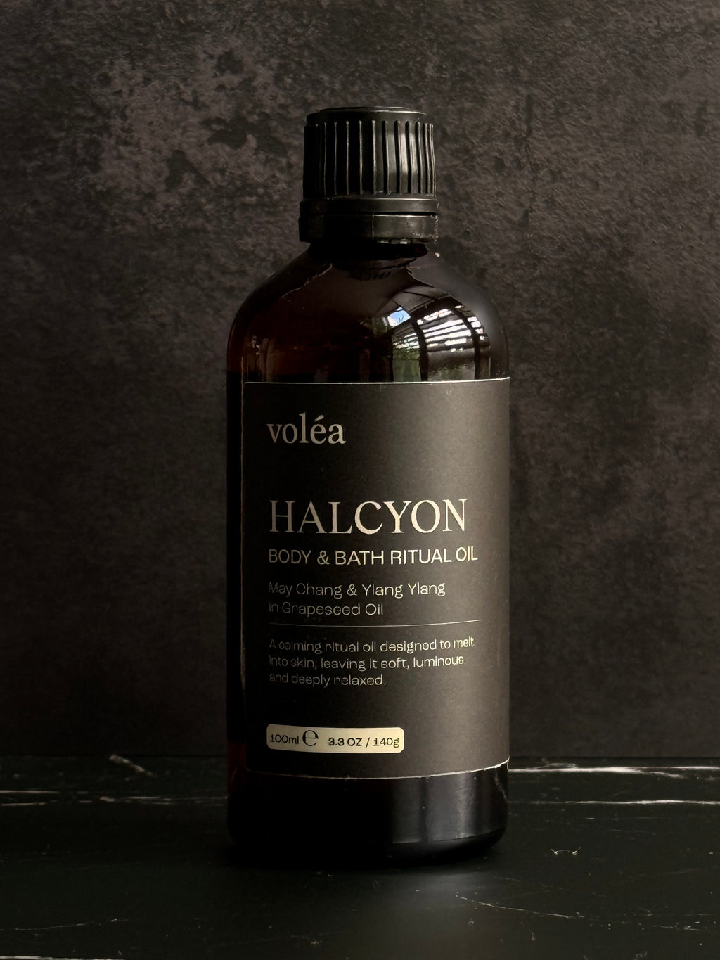 A 330ml bottle of voléa Halcyon Body & Bath Ritual Oil, featuring an amber glass bottle and a black ribbed cap, set against a dark textured background.