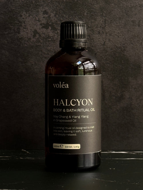 A 330ml bottle of voléa Halcyon Body & Bath Ritual Oil, featuring an amber glass bottle and a black ribbed cap, set against a dark textured background.