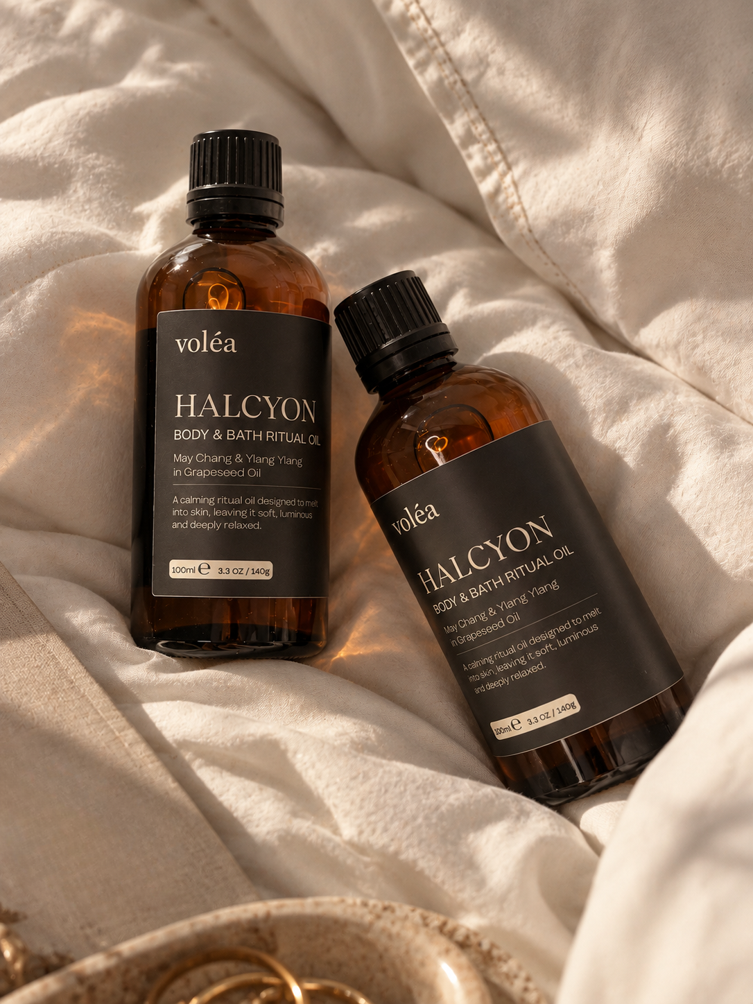 Two amber glass bottles of voléa Halcyon Body & Bath Ritual Oil arranged on draped cream fabric with a decorative bowl edge visible at the bottom.