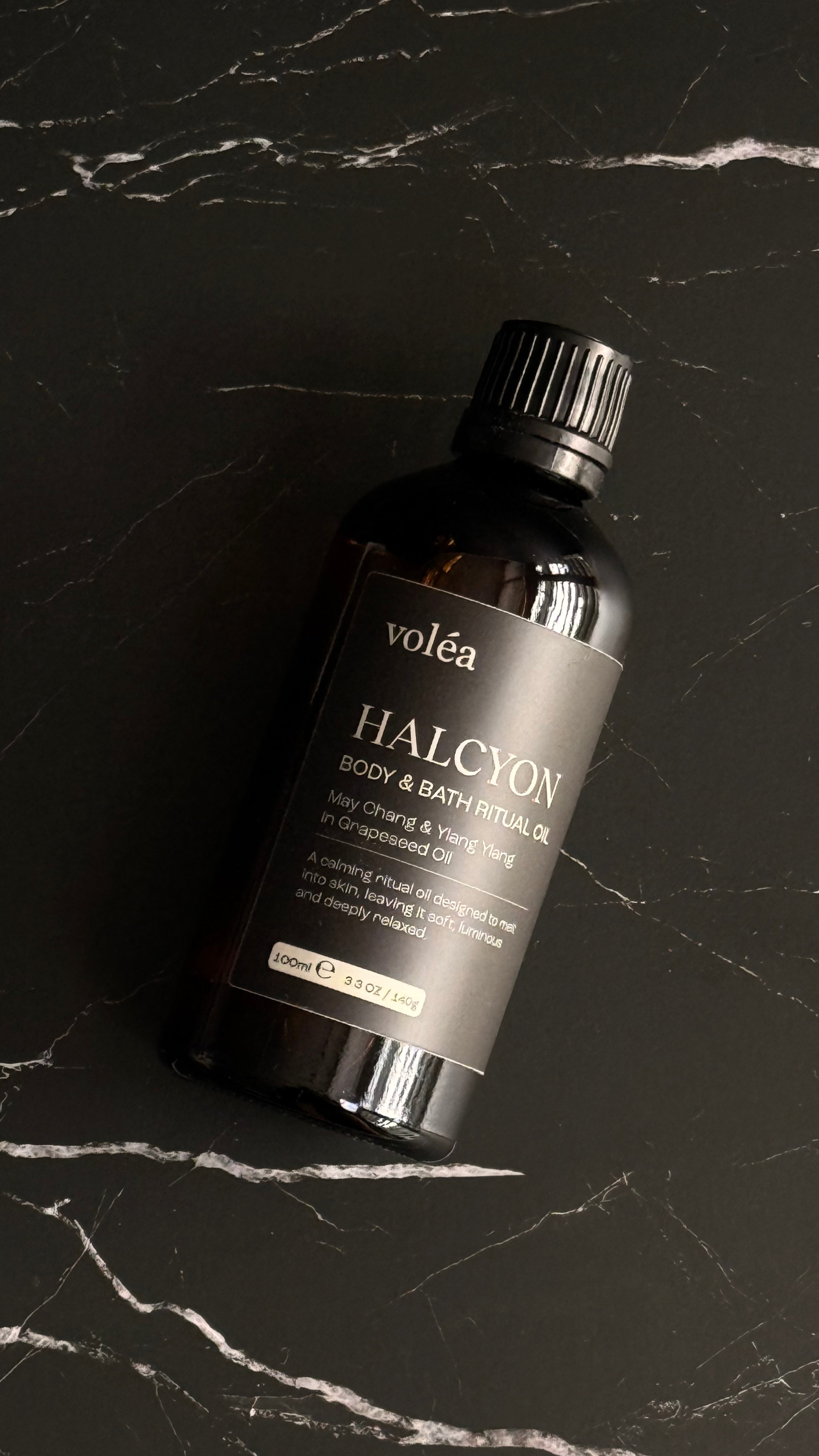 A bottle of Volea Halcyon Body & Bath Rituals resting on a dark marble surface with white veining.