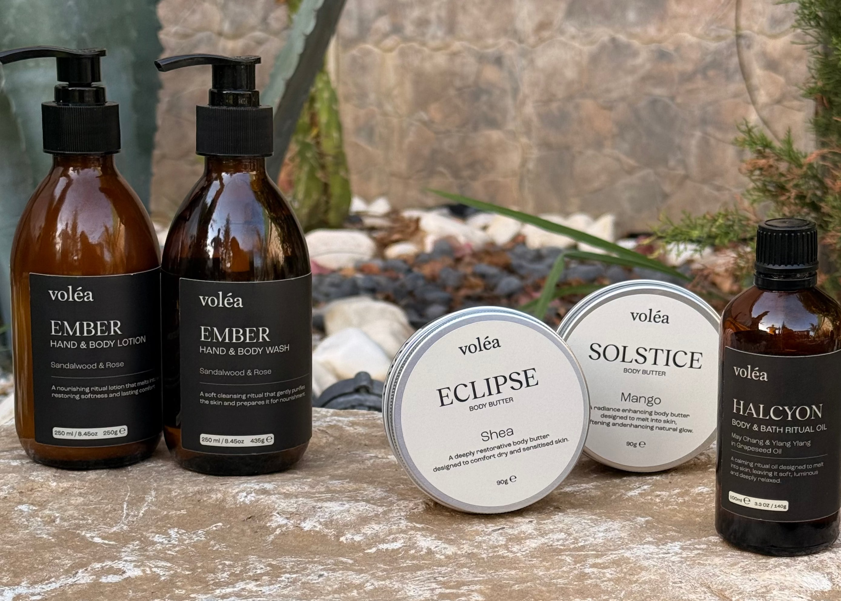 Voléa skincare products arranged on a stone surface outdoors, including two amber pump bottles labeled Ember, two round silver tins labeled Eclipse and Solstice, and one amber bottle labeled Halcyon.