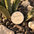 A round tin of voléa Solstice Mango body butter sits on dark river stones between green agave leaves. The label is cream-colored with black text, and the scene is lit by warm natural sunlight.