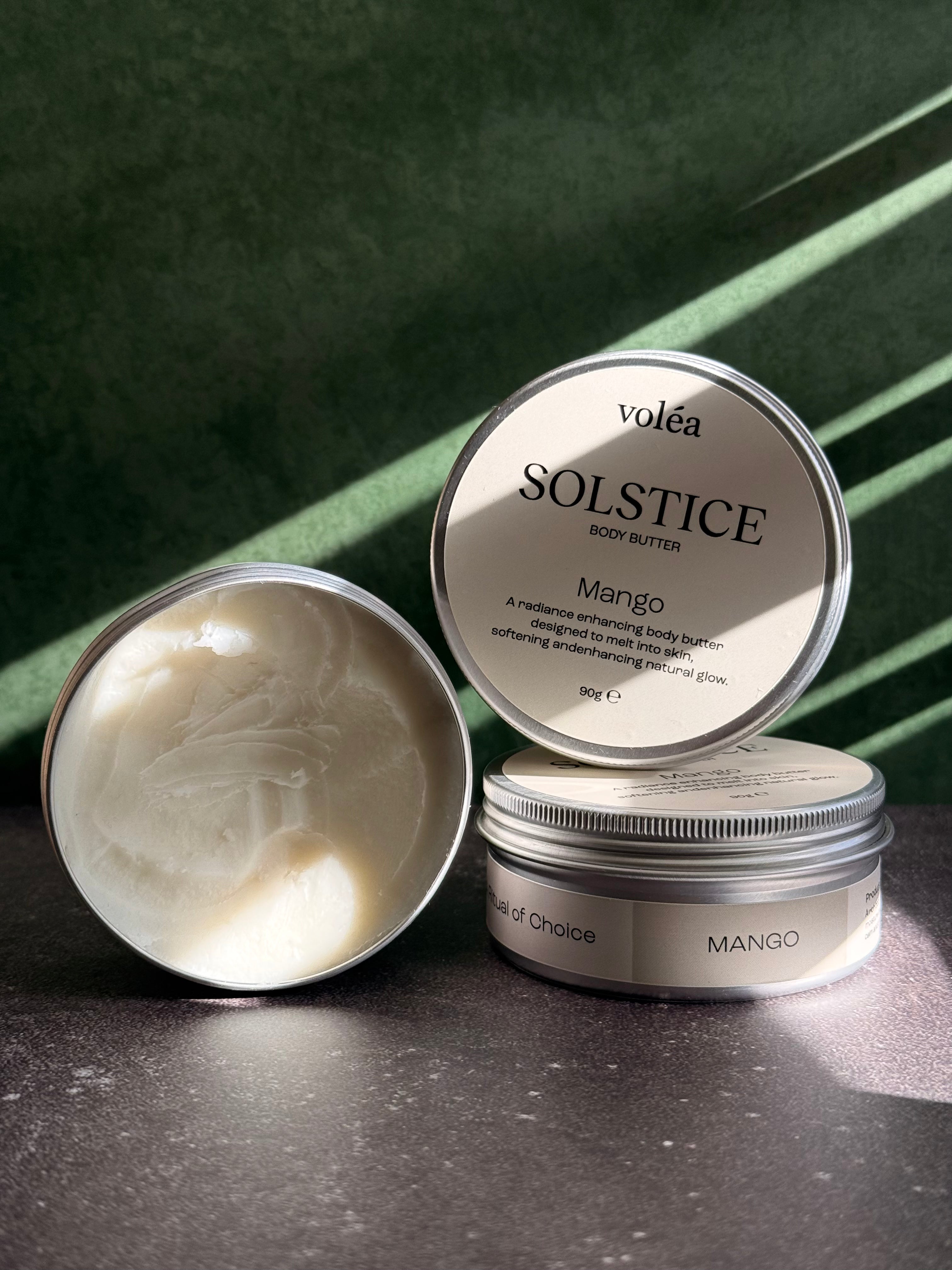 Three round metal jars of Volea Solstice Body Butter in Mango are arranged on a dark surface against a deep green background with diagonal light shadows. One jar is open, showing the creamy off-white body butter inside, while the other two show the labels.