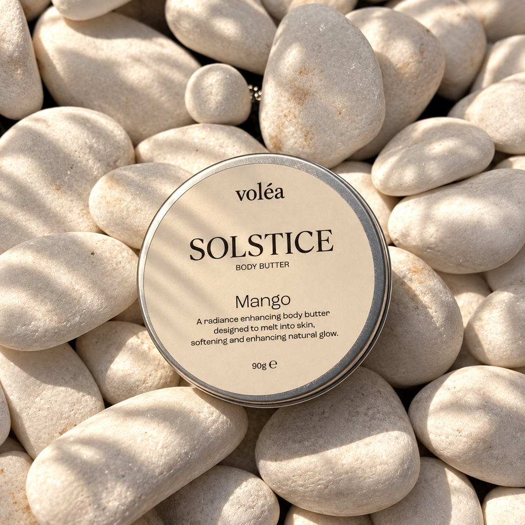 A round tin of voléa SOLSTICE Mango Body Butter lies on a bed of smooth white river stones with soft dappled shadows.