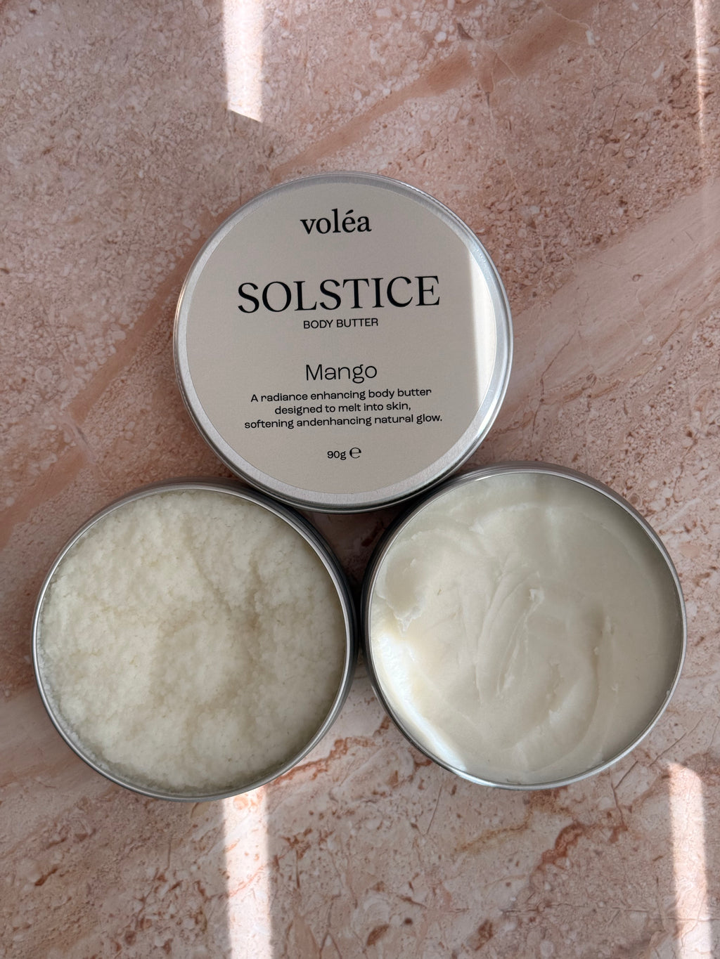 Two open round metal tins of voléa Solstice Body Butter in Mango, showing the white cream inside, placed on a pinkish stone surface with soft sunlight and shadows.