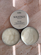 Two open round metal tins of voléa Solstice Body Butter in Mango, showing the white cream inside, placed on a pinkish stone surface with soft sunlight and shadows.