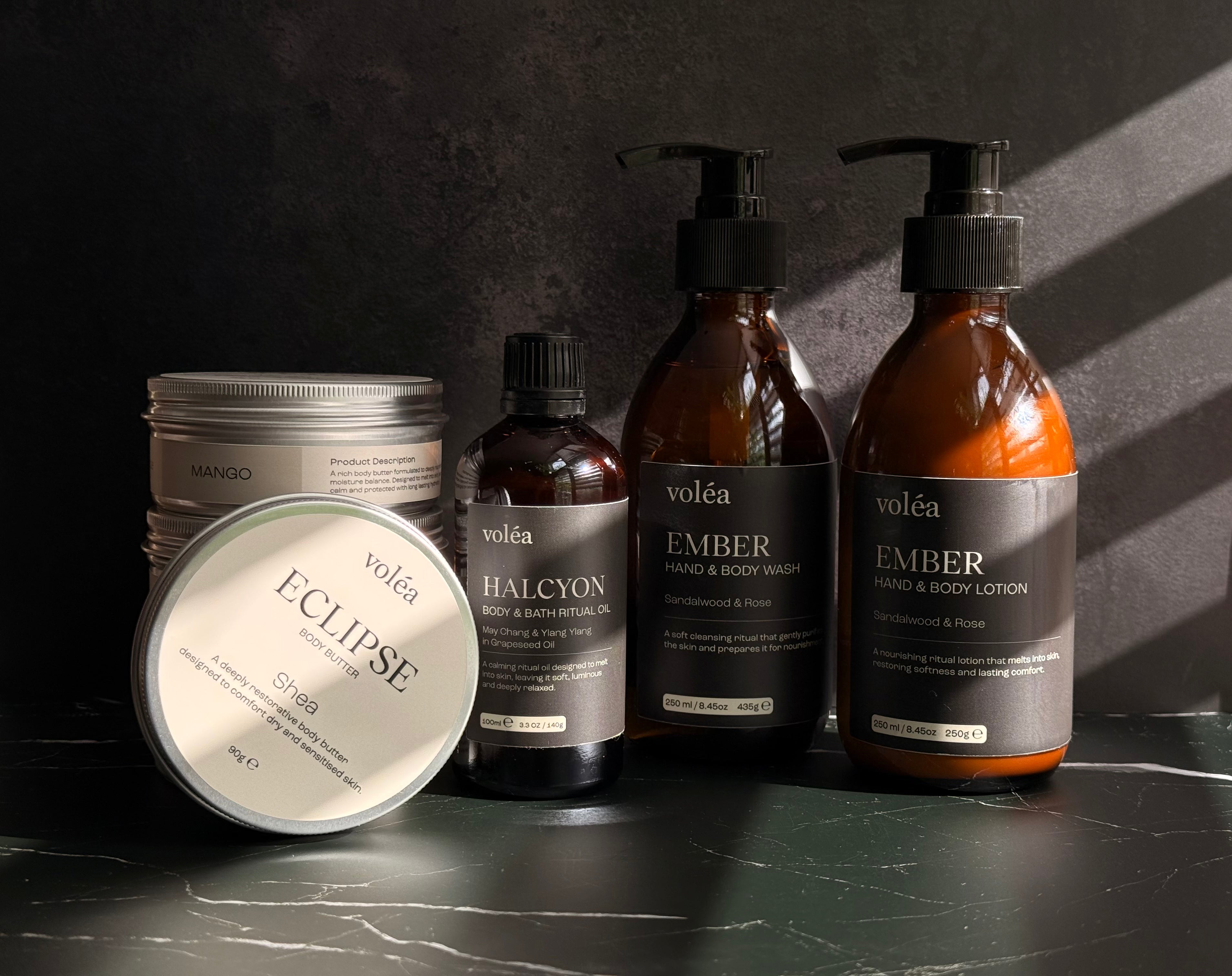 A set of Voléa body and haircare products arranged on a dark marble surface against a dark textured wall, including an Eclipse body butter tin, a Halcyon body wash bottle, an Ember hand & body wash pump bottle, and an Ember hand & body lotion pump bottle.